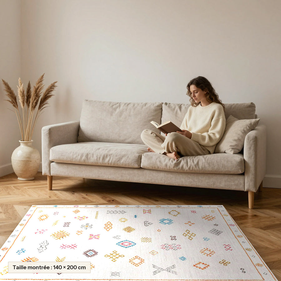 Tapis berbère coloré Tamallat, écru à symboles colorés posé devant un canapé, taille 140 × 200 cm